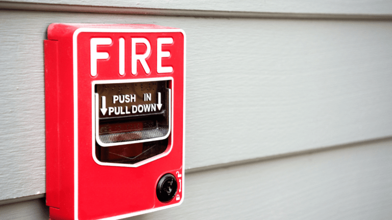 A red fire alarm pull station mounted on a white exterior wall, symbolizing the importance of having a prepared plan of action for pet safety in case of a house fire.